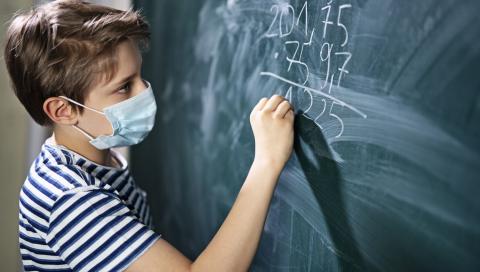 Young boy with mask does math problem on blackboard